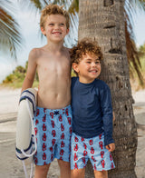Baby boy wearing navy long sleeve rash guard by RuffleButts + RuggedButts, smiling outdoors in blue sun protection swimwear.