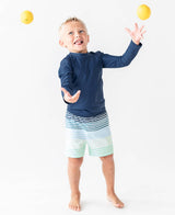 Baby boy wearing navy long sleeve rash guard by RuffleButts + RuggedButts, blue sun protection swimwear for infants.