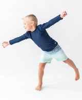 Baby boy wearing navy long sleeve rash guard by RuffleButts + RuggedButts, showing the blue protective swimwear for infants.