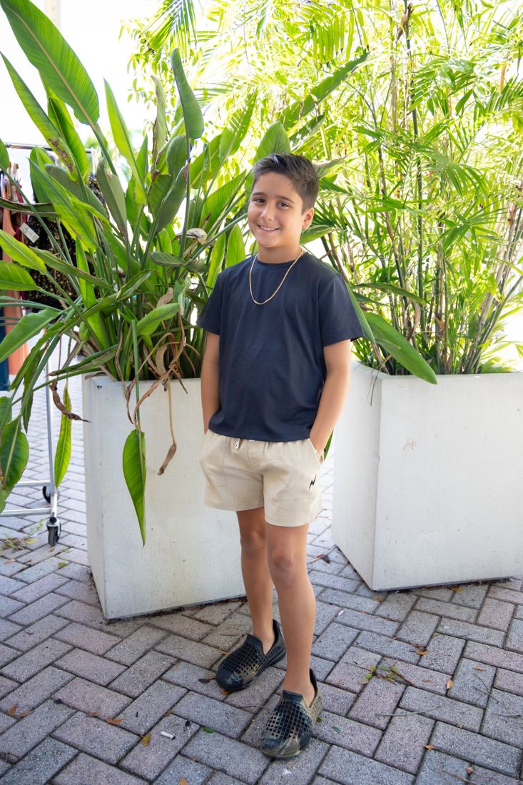 Boy in a navy tee and khaki shorts standing by tropical plants