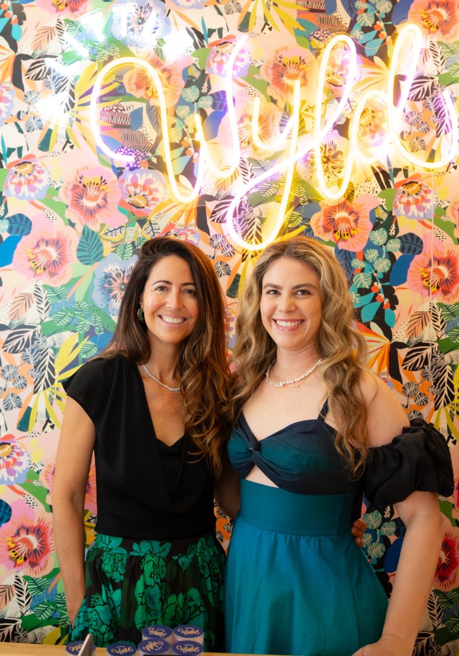 A Little Wyld founders Amanda Perna and Skye Dyer smiling under the neon Wyld sign in their Delray Beach boutique
