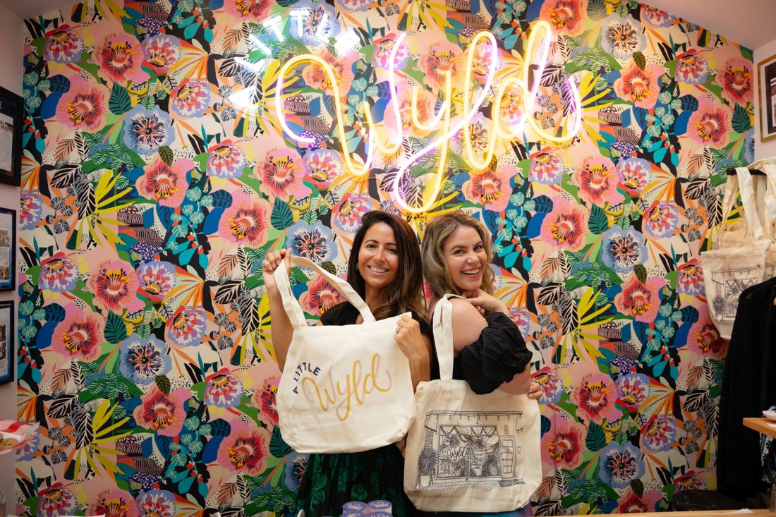 Amanda and Skye holding A Little Wyld tote bags in front of the floral wallpaper